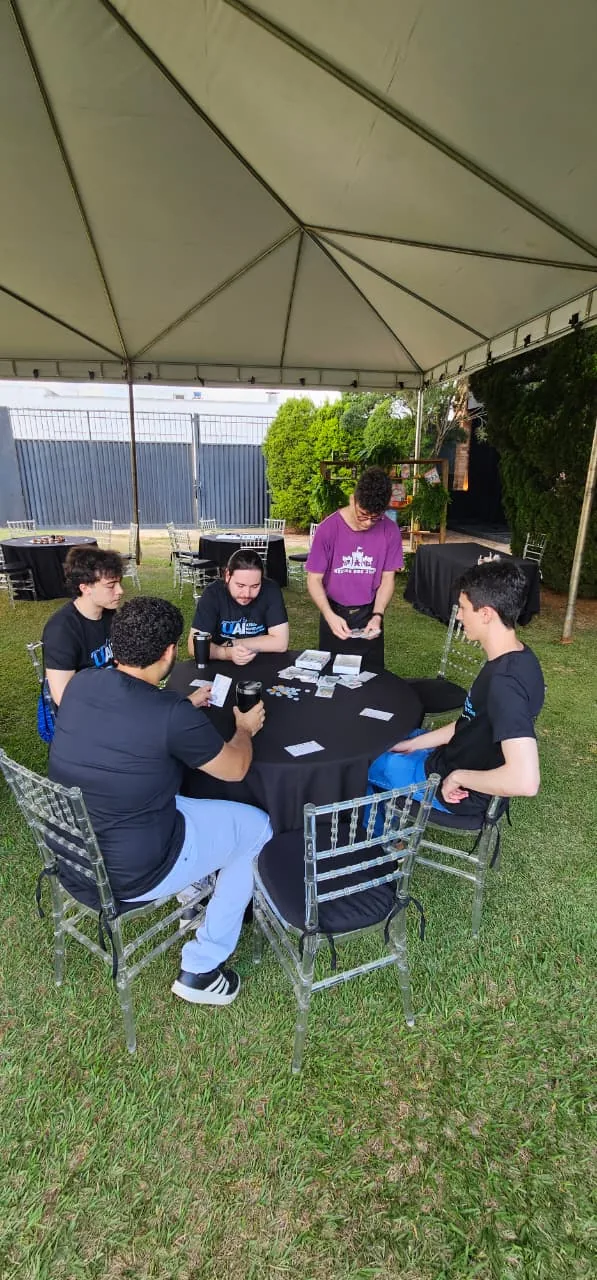 Grupo reunido jogando um jogo de tabuleiro numa mesa redonda ao ar livre, com um monitor da Mestre ensinando em pé