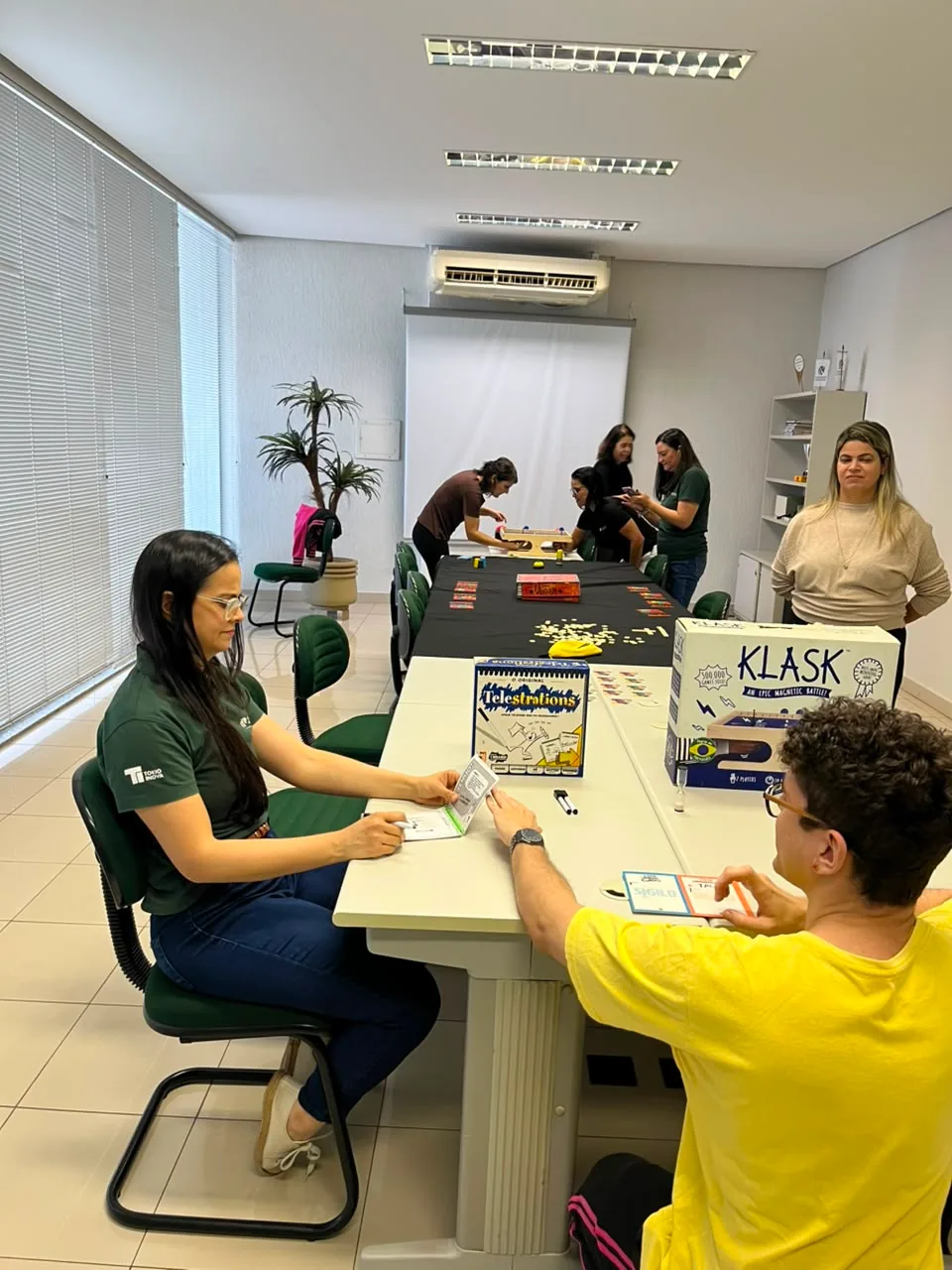 Colaboradores jogando jogos de tabuleiro numa sala de reunião, com caixas de jogos sobre a mesa e um monitor acompanhando