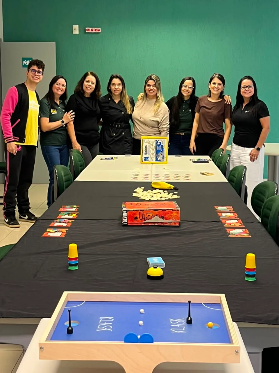 Equipe de uma empresa reunida e sorrindo atrás da mesa preparada com vários jogos para a confraternização