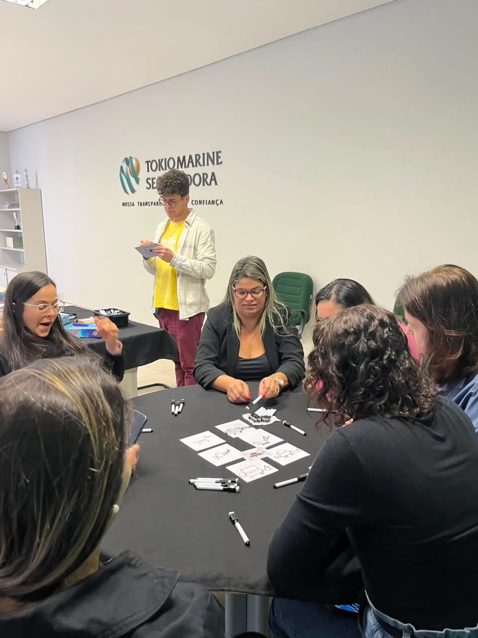 Colaboradores ao redor de uma mesa numa dinâmica de desenho, com cartas e canetas espalhadas, e um monitor conduzindo em pé