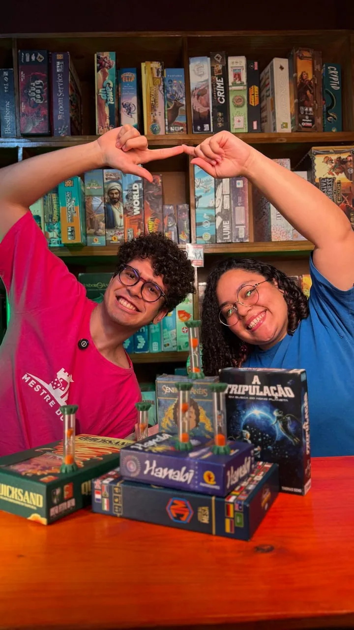 Dois monitores da Mestre dos Jogos fazendo um coração com as mãos, em frente à estante de jogos