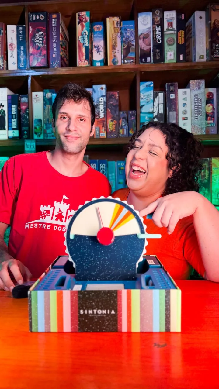Dois monitores da casa sorrindo atrás do jogo Sintonia, com a estante de jogos ao fundo