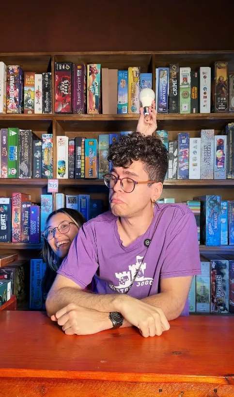 Dois monitores brincando atrás do balcão, um equilibrando uma lâmpada sobre a cabeça, com a estante de jogos ao fundo
