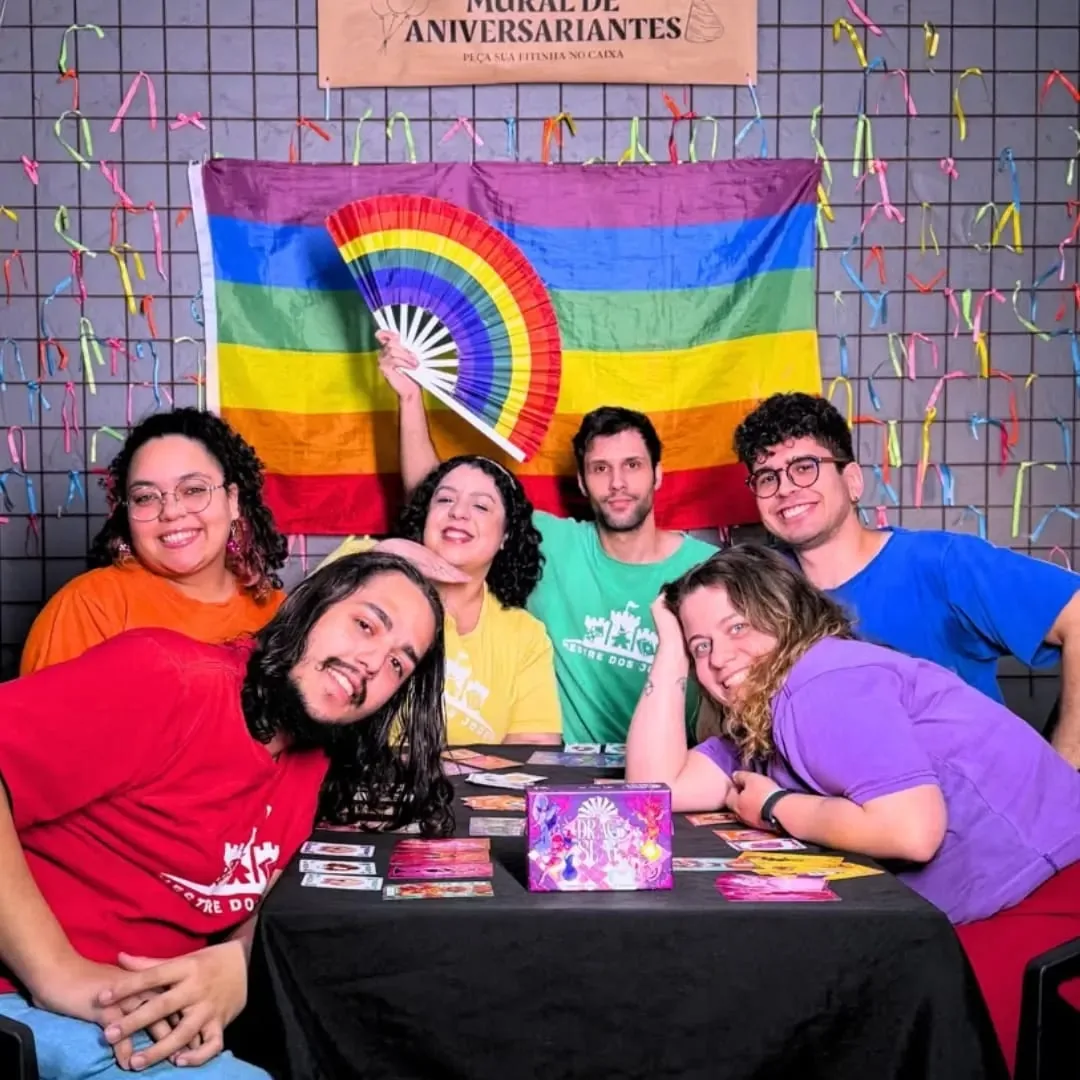 A equipe de monitores da Mestre dos Jogos reunida à mesa, sorrindo, com uma bandeira de arco-íris ao fundo