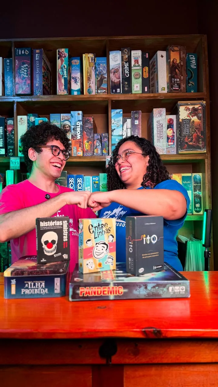 Dois monitores da casa sorrindo com jogos na mesa, em frente à estante