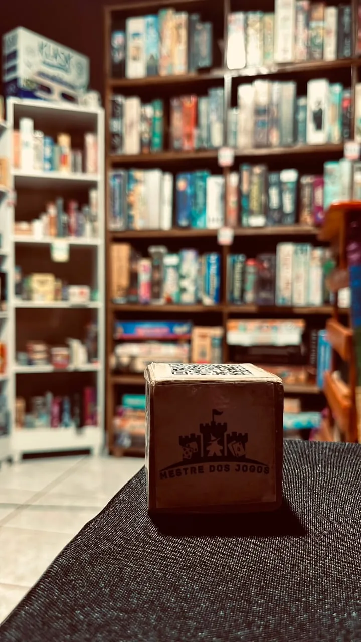 Cubo de madeira com o brasão da Mestre dos Jogos e um QR code, sobre a mesa, com a estante de jogos ao fundo