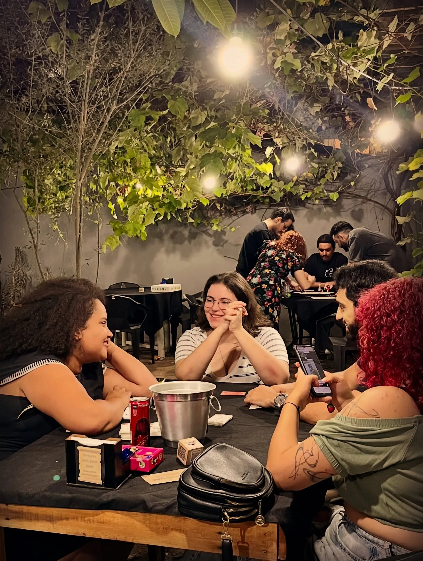 Amigos rindo ao redor da mesa sob a pérgula de plantas e luzinhas, no salão da Mestre dos Jogos
