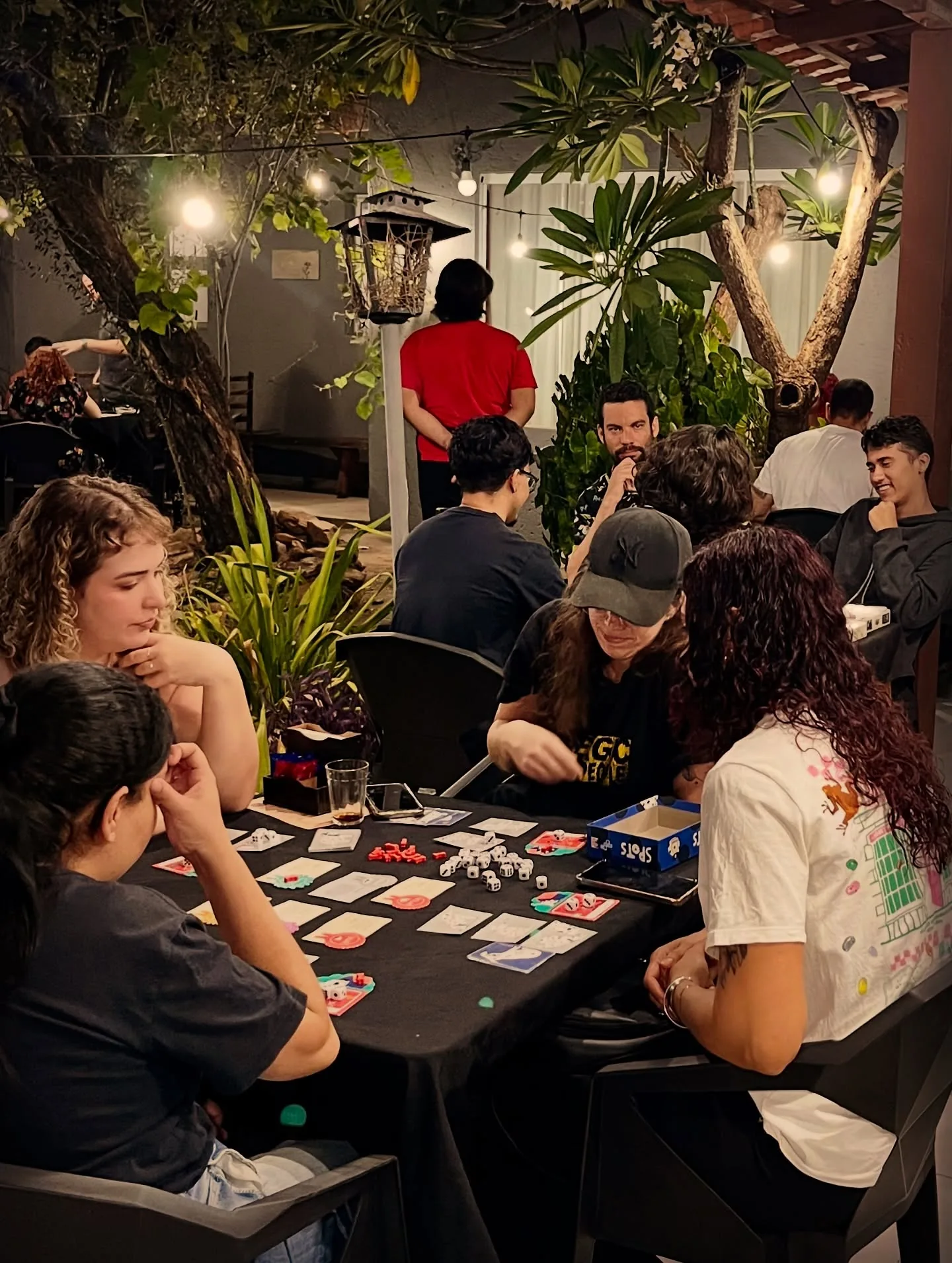Mesas cheias de gente jogando à noite, entre árvores e luzes do jardim
