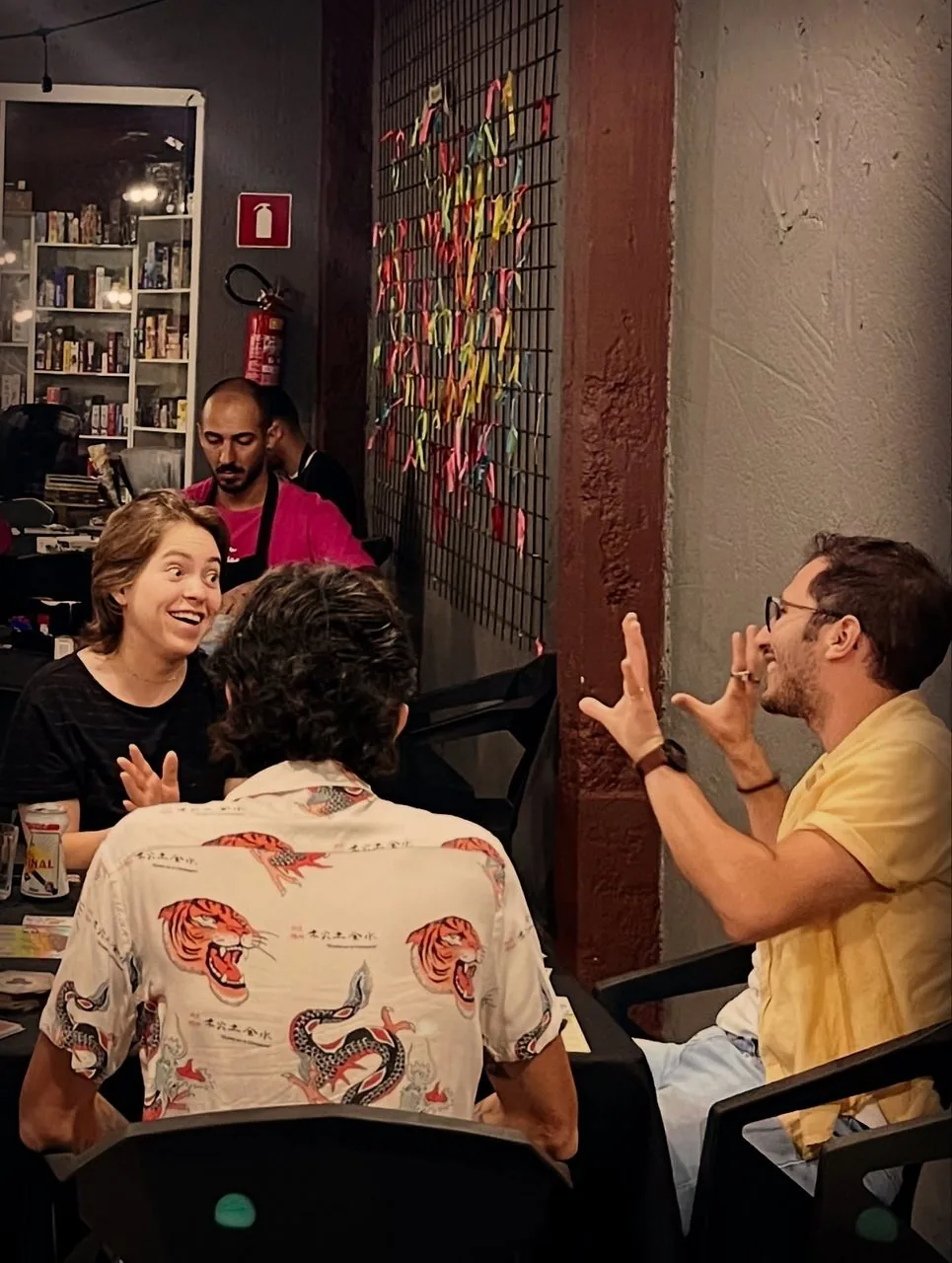 Clientes conversando ao lado da estante cheia de jogos, com um monitor ao fundo