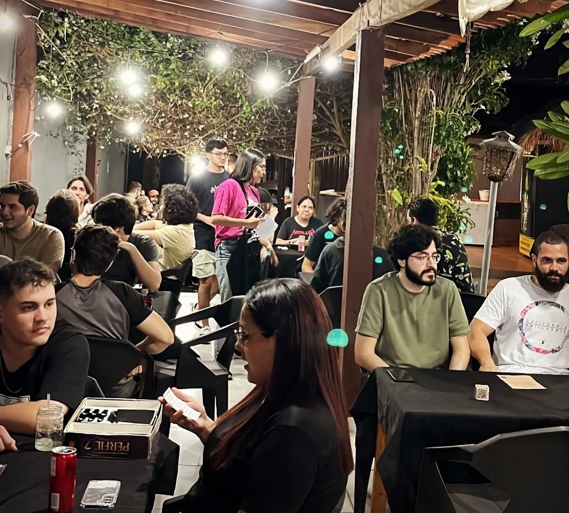 Salão cheio sob a pérgula de luzinhas, com várias mesas de gente jogando à noite