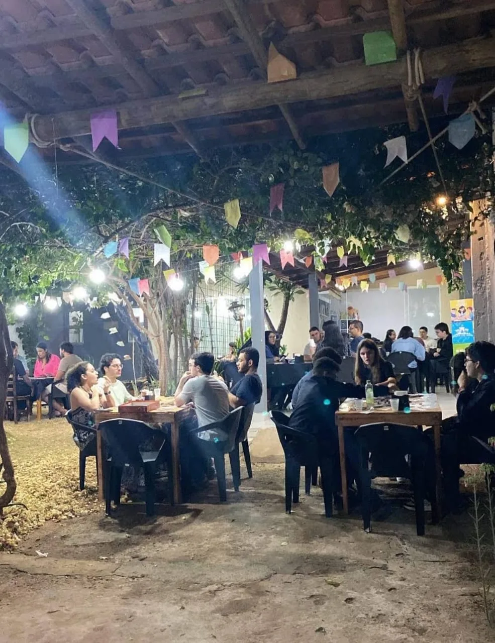 Salão lotado à noite, com várias mesas de gente jogando sob as bandeirinhas e luzes do jardim