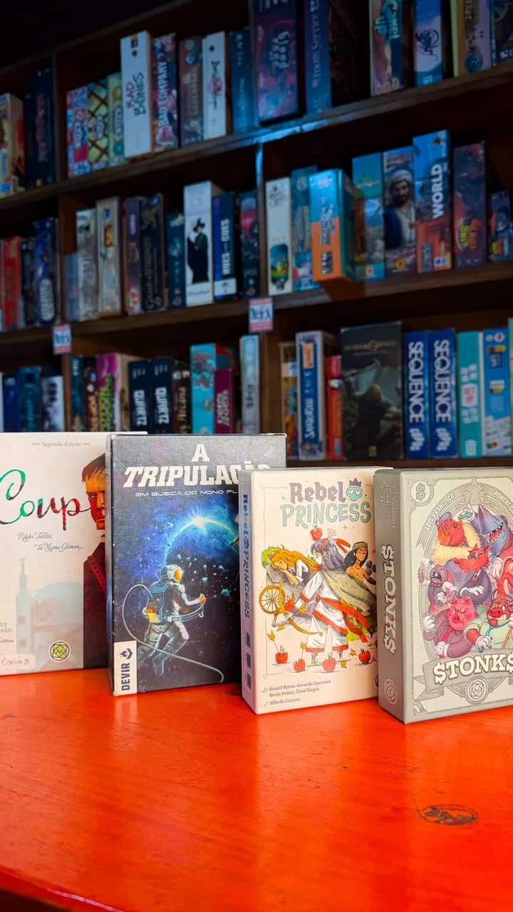 Jogos de cartas da casa alinhados sobre a mesa, com a estante de tabuleiros ao fundo