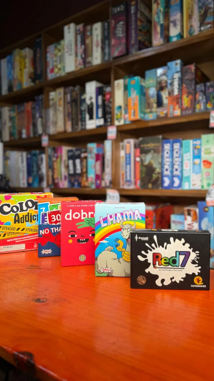Caixas de jogos de cartas em pé sobre a mesa de madeira, com a estante ao fundo